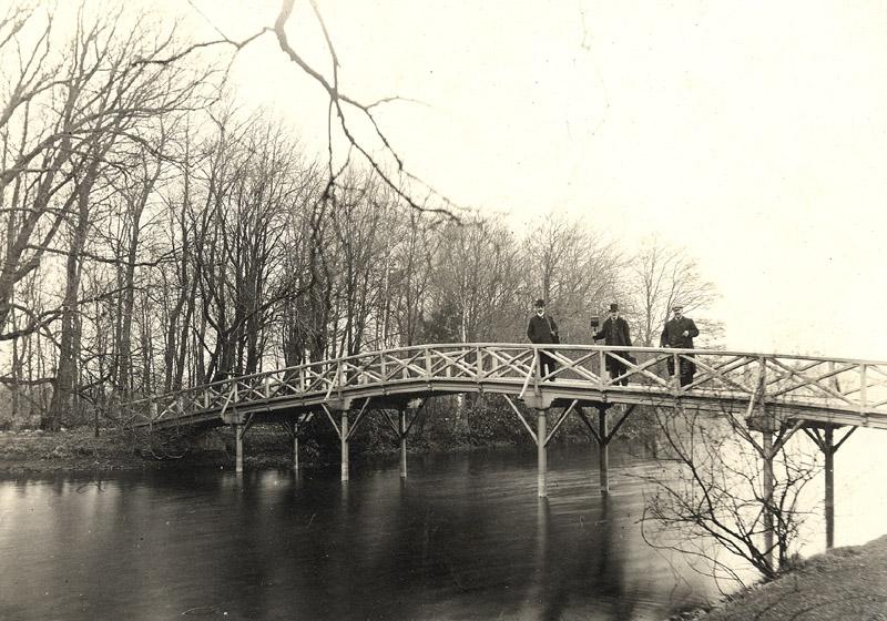 Historical black and white image of three men standing on a bridge over a lake at Hartsholme Country Park with lots of trees in the background