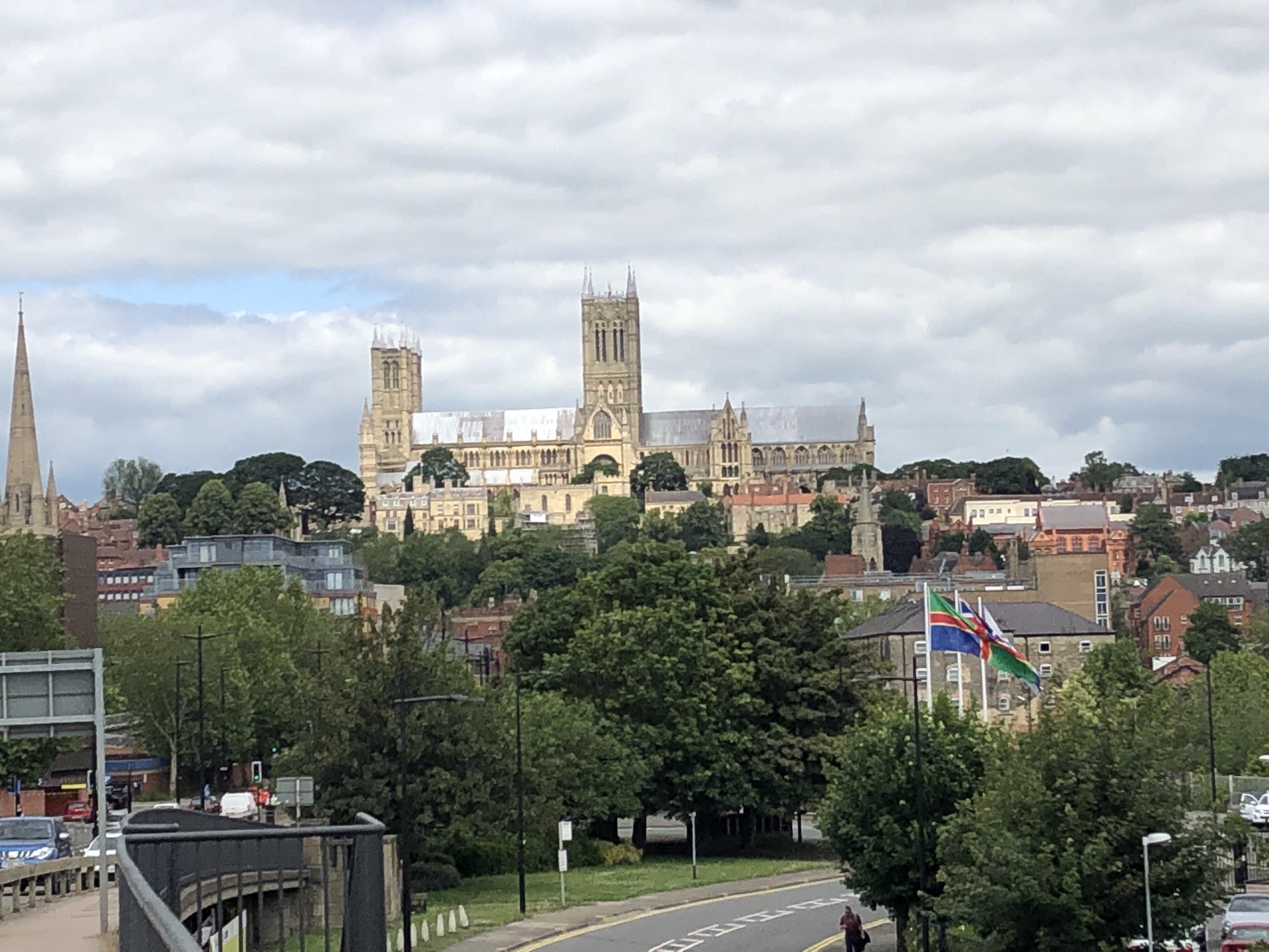 Lincoln Cathedral taken from the south of the city. The Cathedral is high up with lots of trees, and other buildings, in front of it and there are some Lincolnshire flags waving in the breeze.