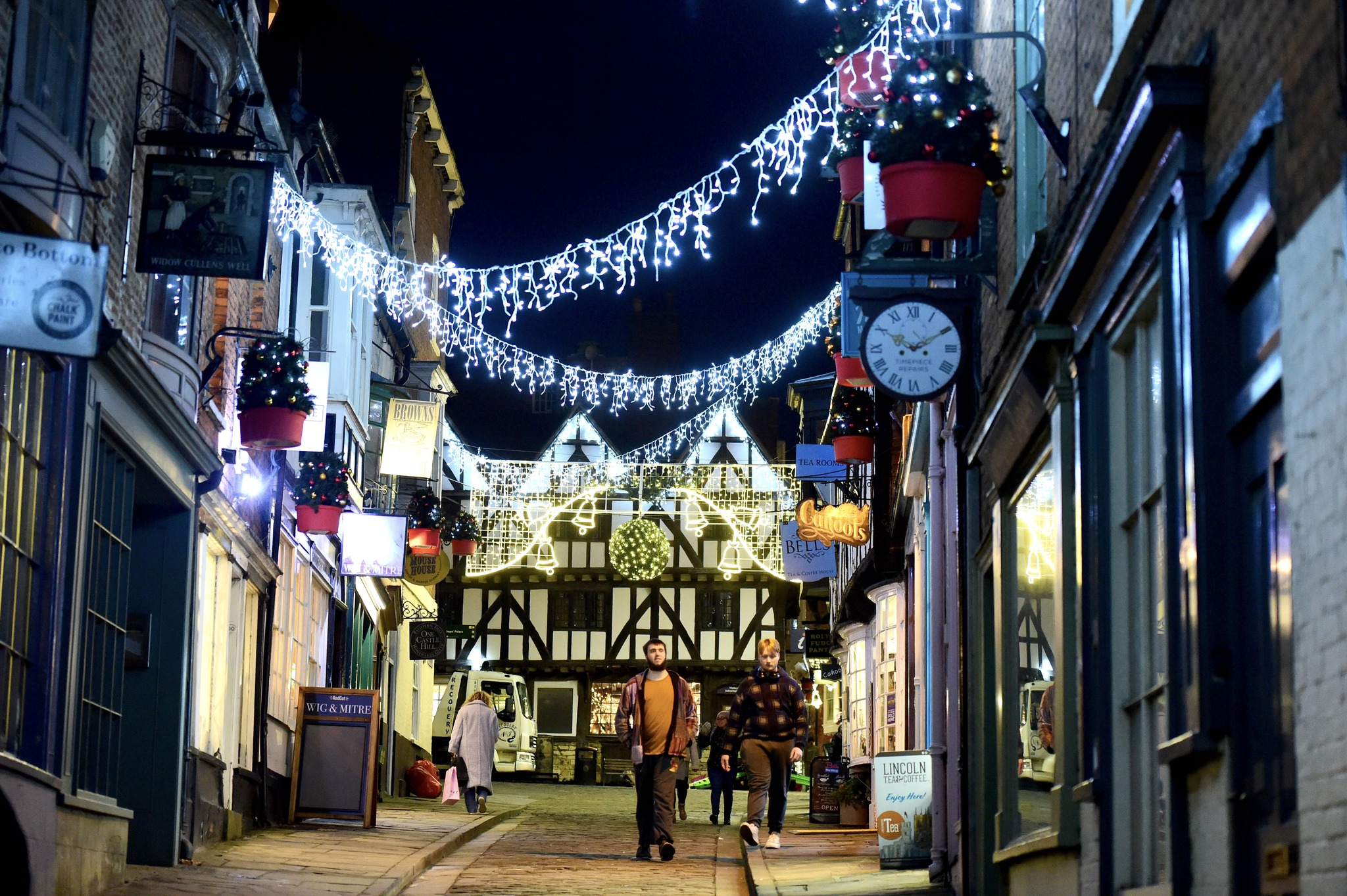 Christmas lights hanging from shops at the top of Lincoln high street with a white male walking towards the camera.