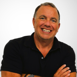 Crispin wears a black polo shirt and grins at the camera with his arms crossed.