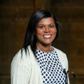 Mel wears a polka dot black and white top and white cardigan, as she smiles enthusiastically at the camera.