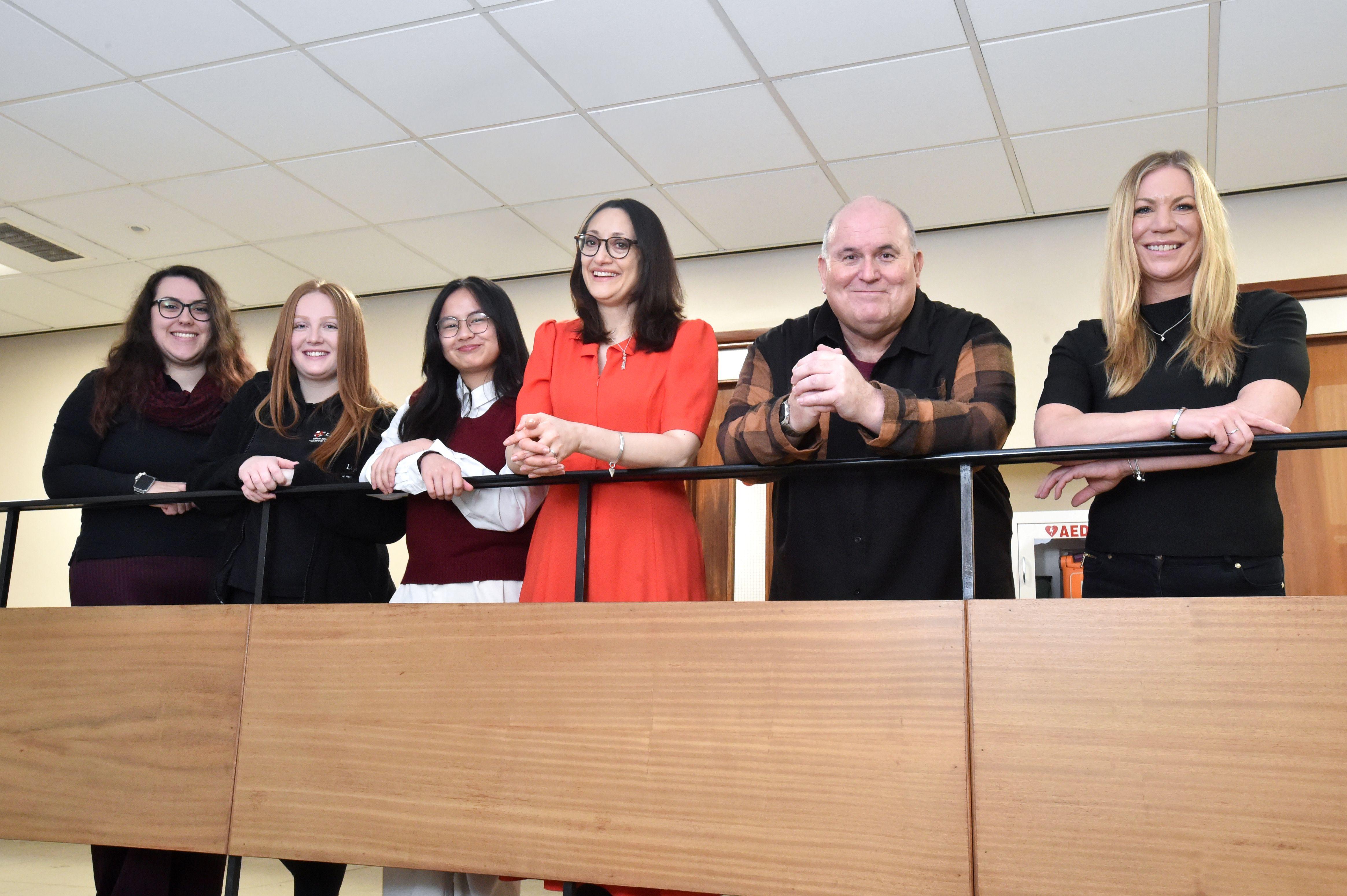 A row of five women and one man leaning on a banister of a stair well. All are dressed in black apart from the Portfolio Holder who is wearing red.