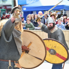 Two Vikings facing each other in the arena hold wooden shields and swing their swords up high.