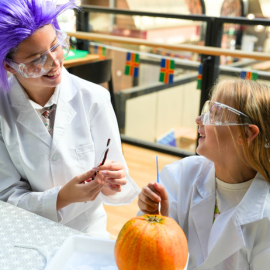 A little girl dressed as a scientist looks up smiling at a woman dressed as a mad scientist. The little girl is painting a pumpkin.
