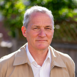 Gregory Hughes stands in a sunny outdoor setting, looking at the camera with trees in the background.
