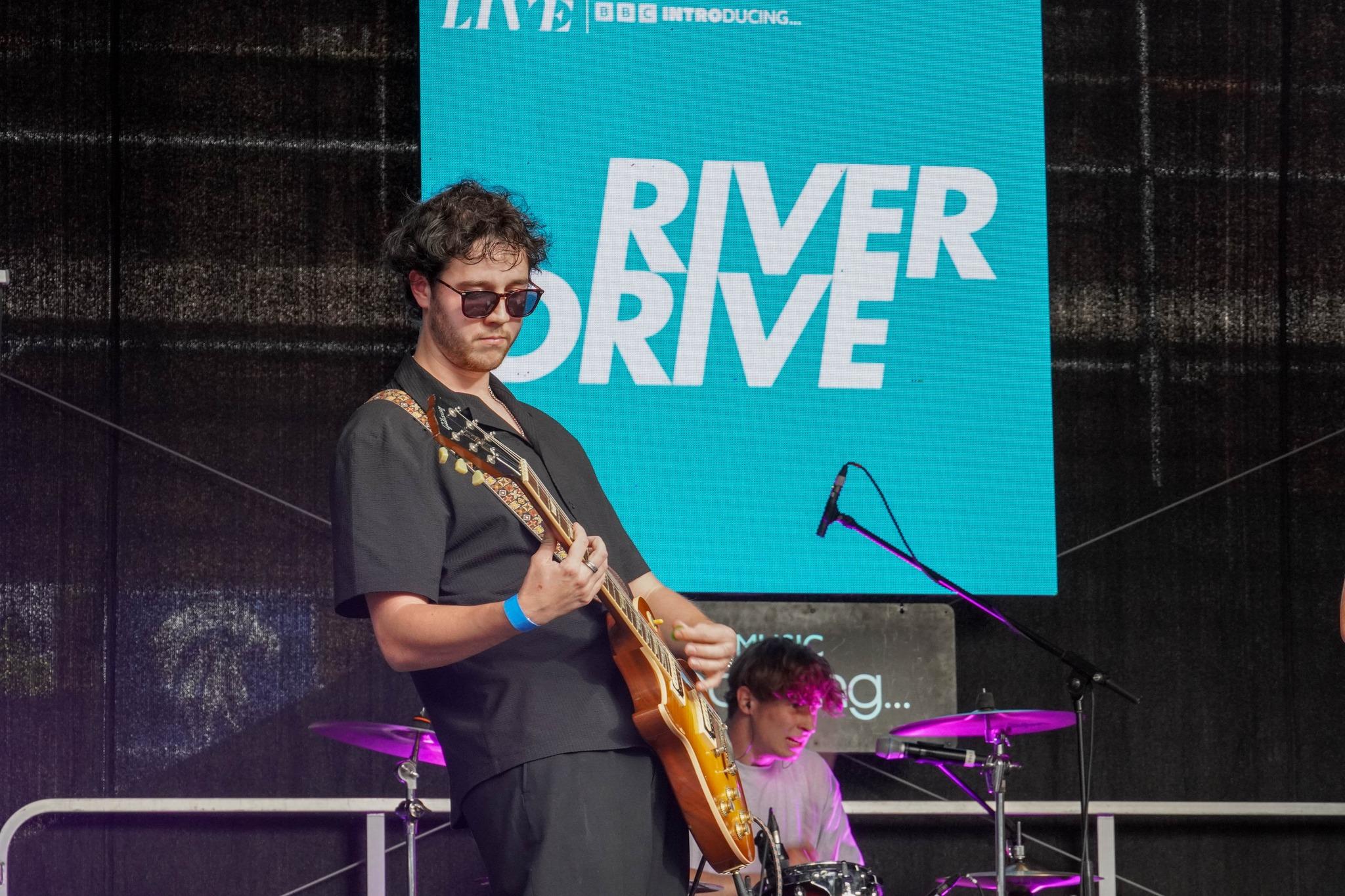 A man wearing sunglasses and playing the guitar on stage with a blue screen behind him that reads River Drive.