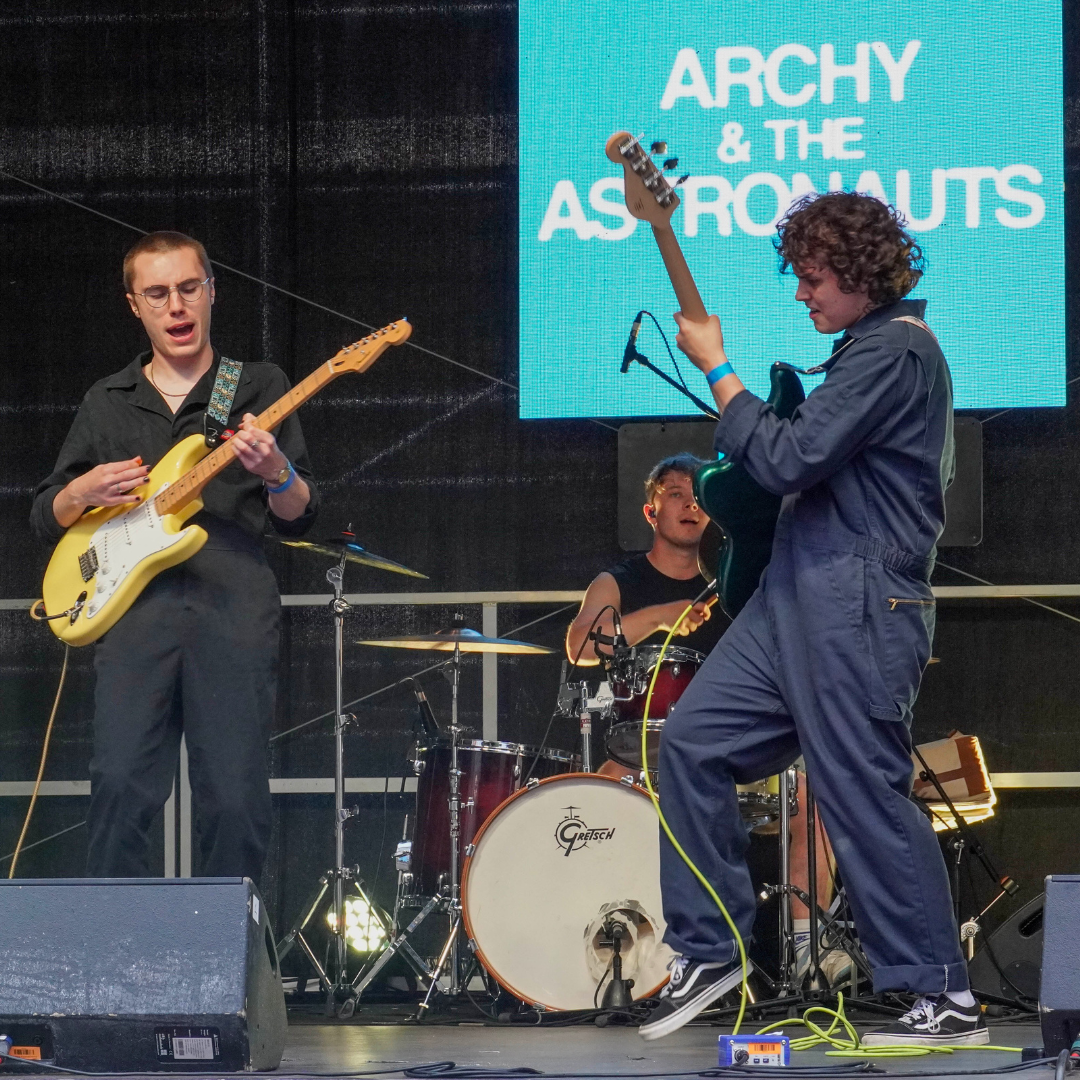 Members of Archy and the Astronauts are live on stage; there are two at the front playing guitars and a drummer in the background.