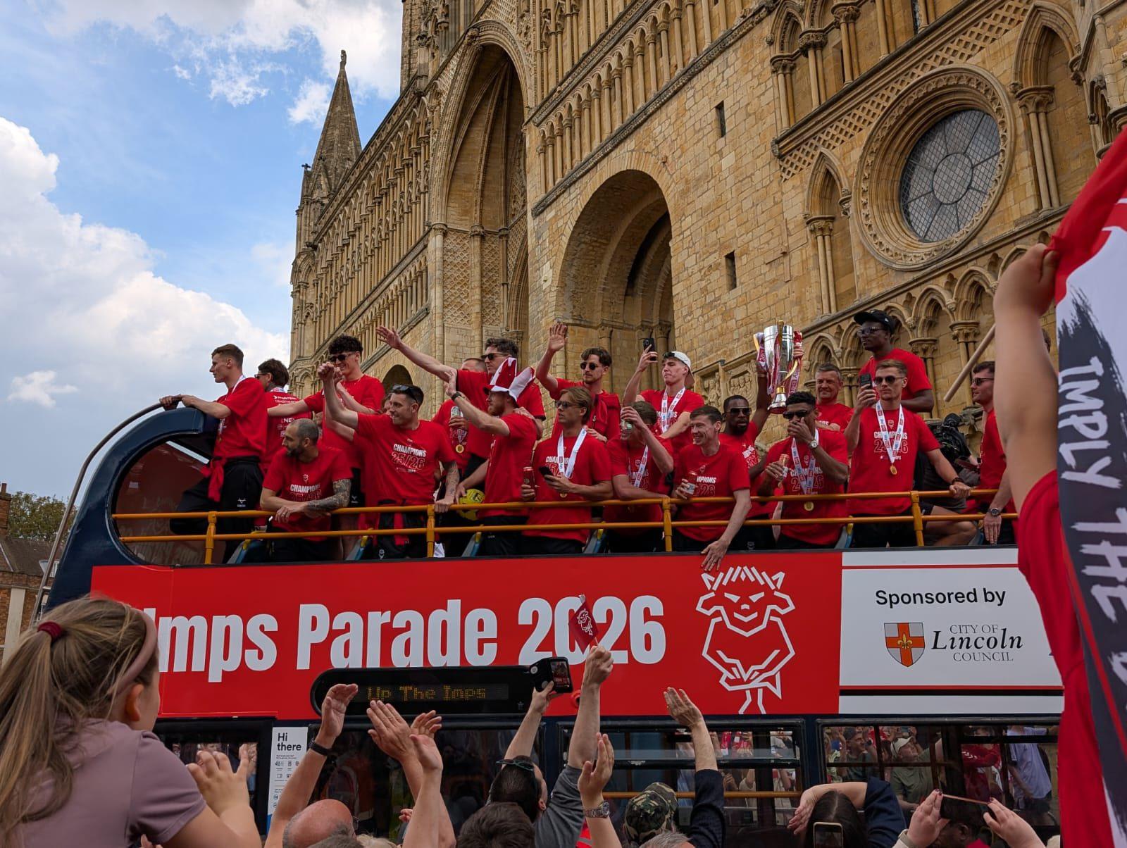 Lincoln City FC players on an open top bus with their arms in the air in front of Lincoln Cathedral with many crowds below.