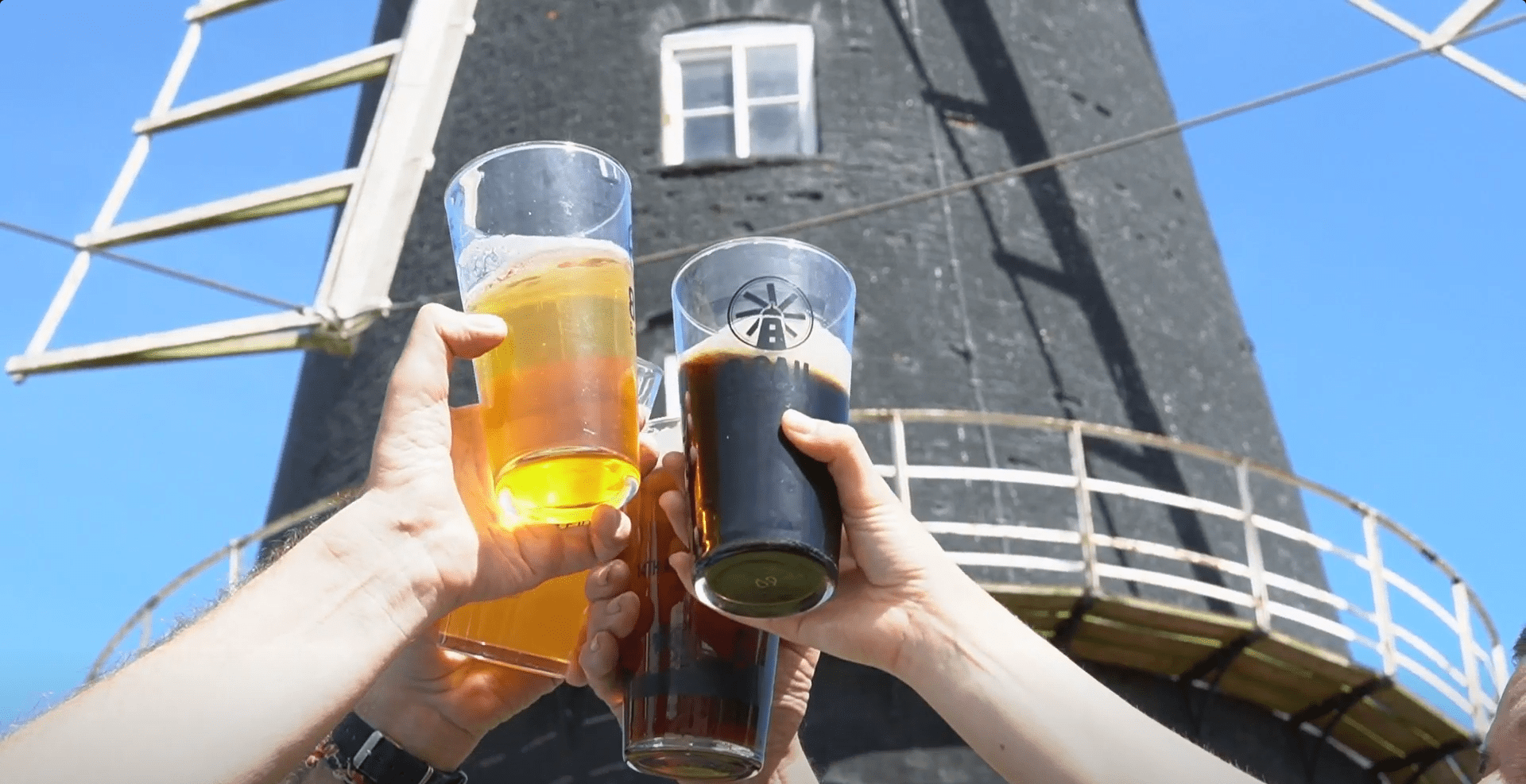 People are holding up beers to say cheers in front of a windmill