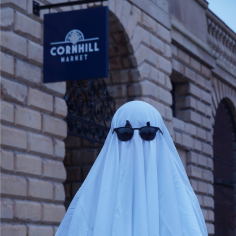 A person in a sheet ghost costume wears sunglasses outside Cornhill Market's entrance.