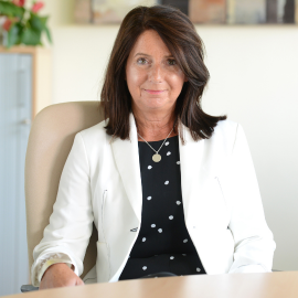 Angela sits smiling at the camera at her desk, wearing a black shirt and white blazer.