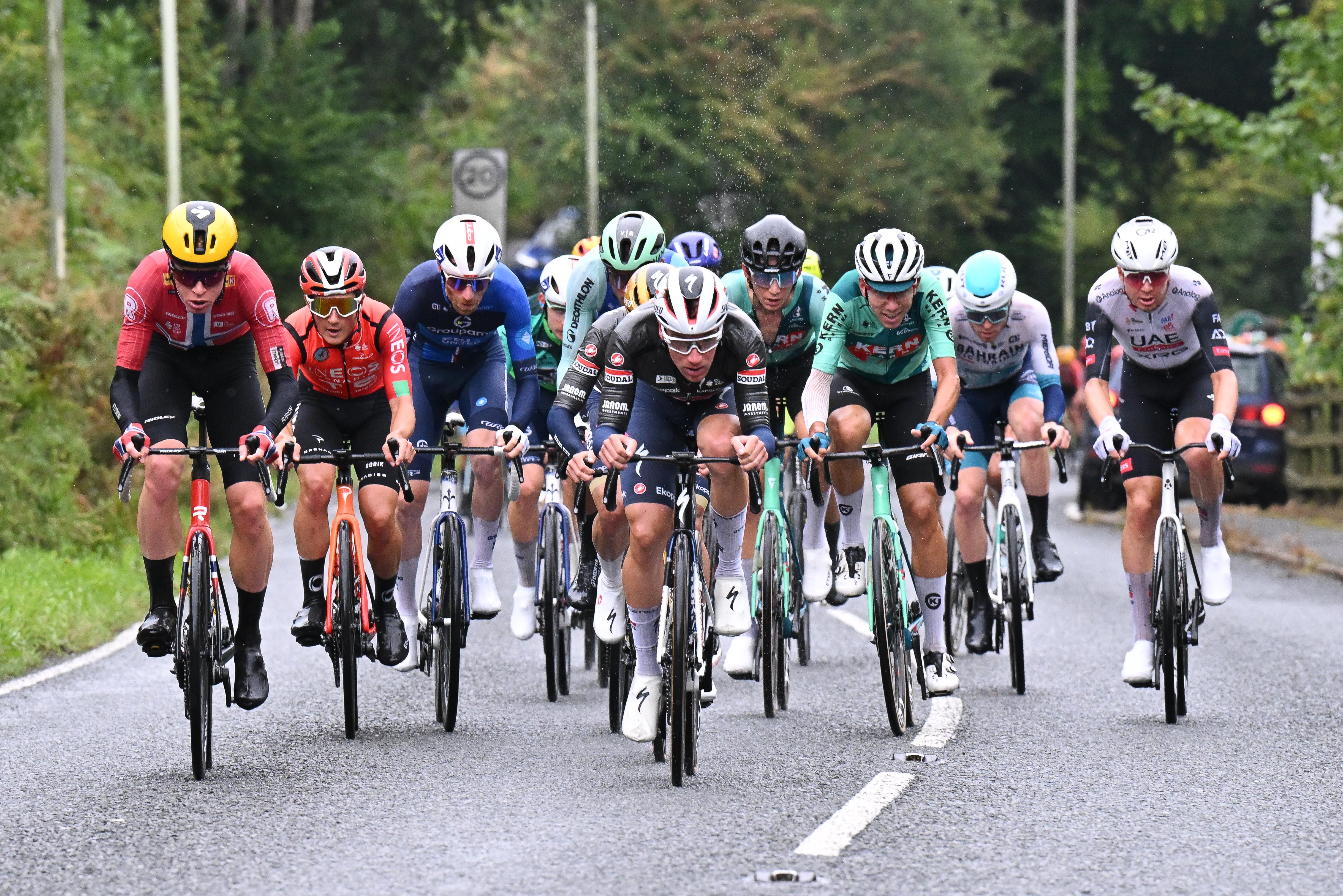 A cluster of professional male cyclists riding their bikes as part of the Tour of Britain 2025 on a main road with trees either side.