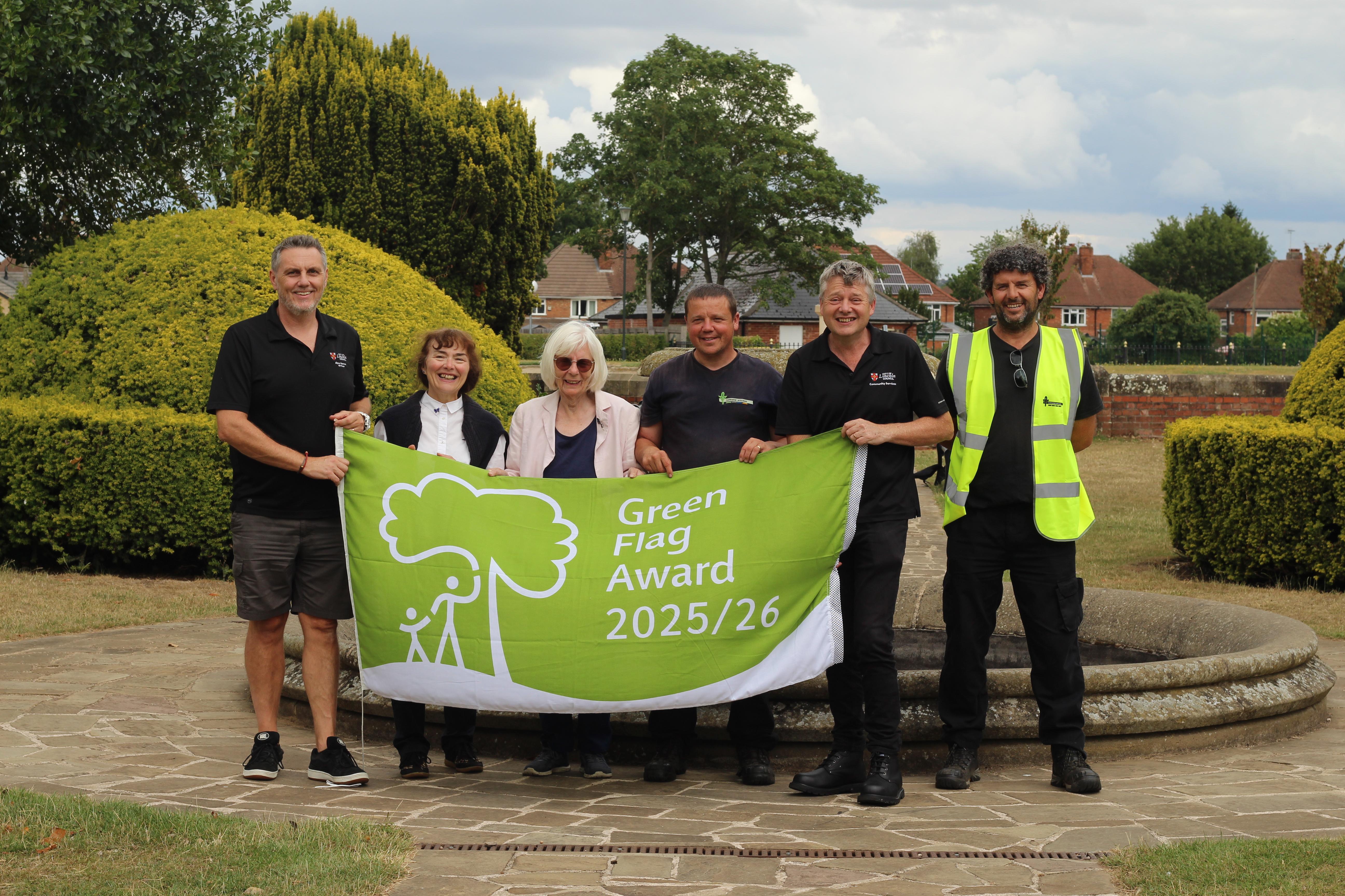 A man, two women and three men stood in a line all holding a large green flag that says Green Flag Award 25/26