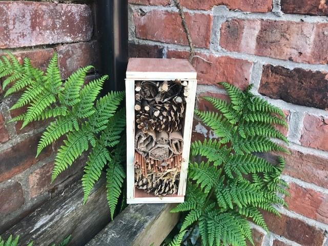 A wooden bug box hotel with leaves all around it, in front of a brick wall.