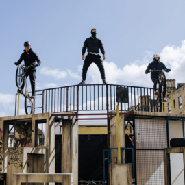 Three stunt men/performers are on the top of on an urban structure, two of the men are on bikes.