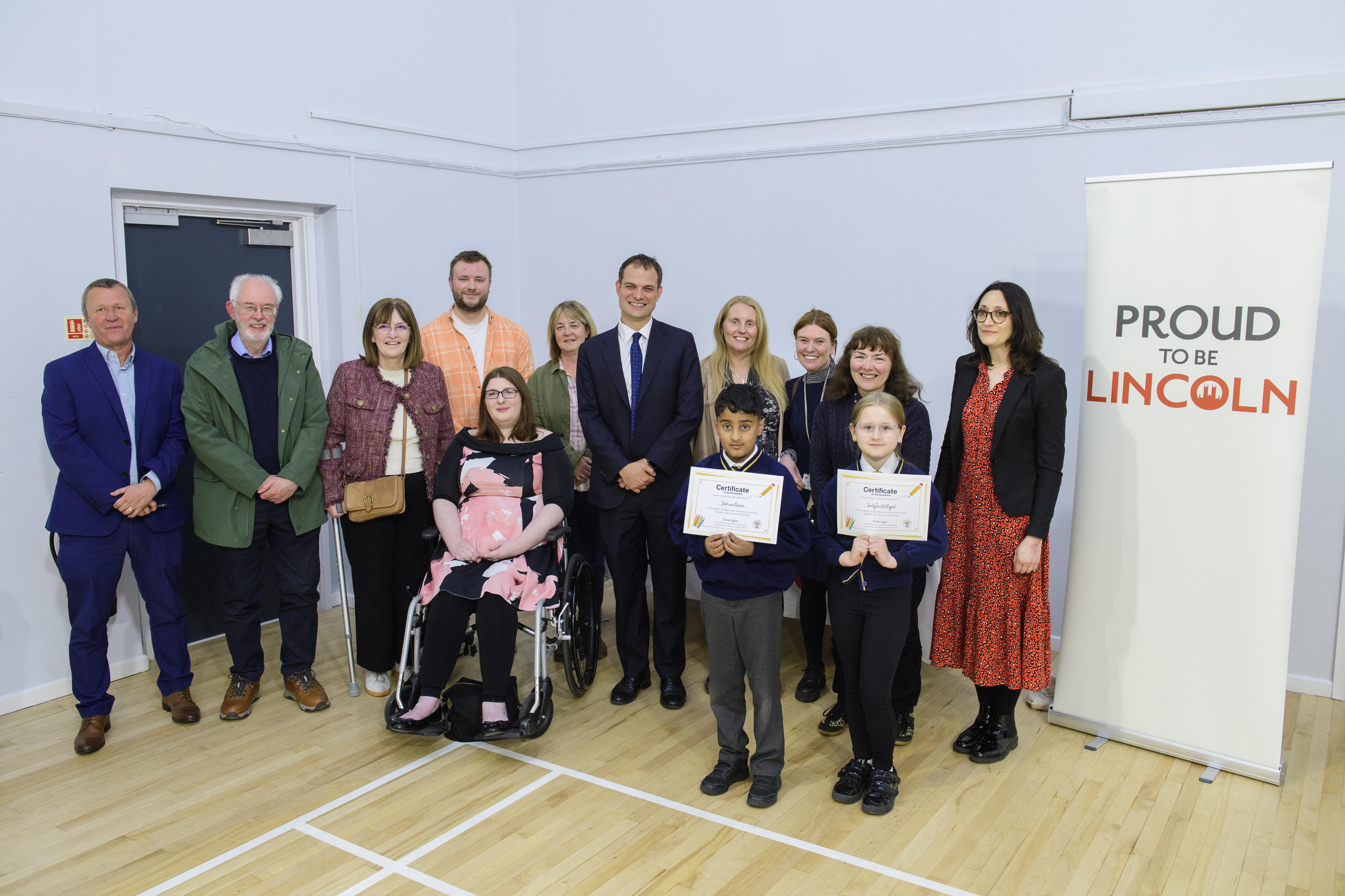 Councillors, staff, and residents posed for a photo next to a Proud to be Lincoln banner with two school children holding certificates.