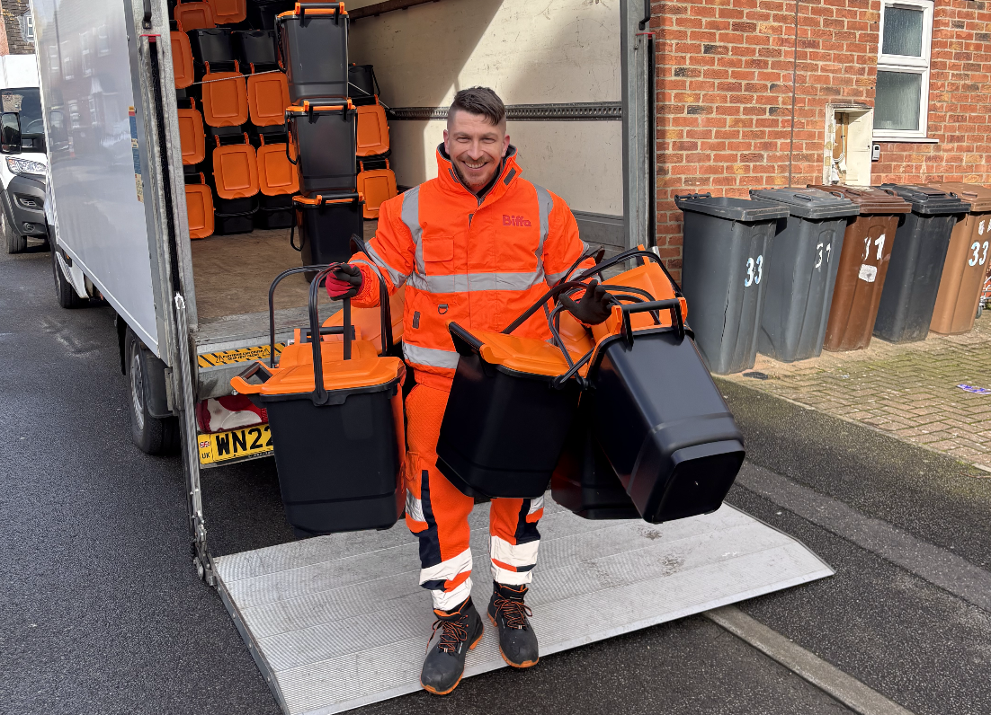 A man in a hi-vis orange uniform stood at the end of a lorry carrying three outdoor food waste caddies on each arm while grinning.