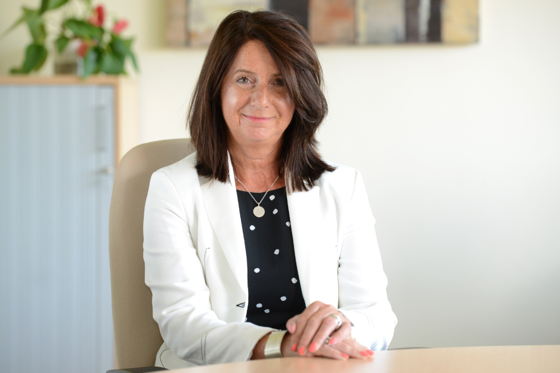 Angela Andrews, wearing a white blazer, sits behind a desk in an office smiling at the camera with a plant in the background.