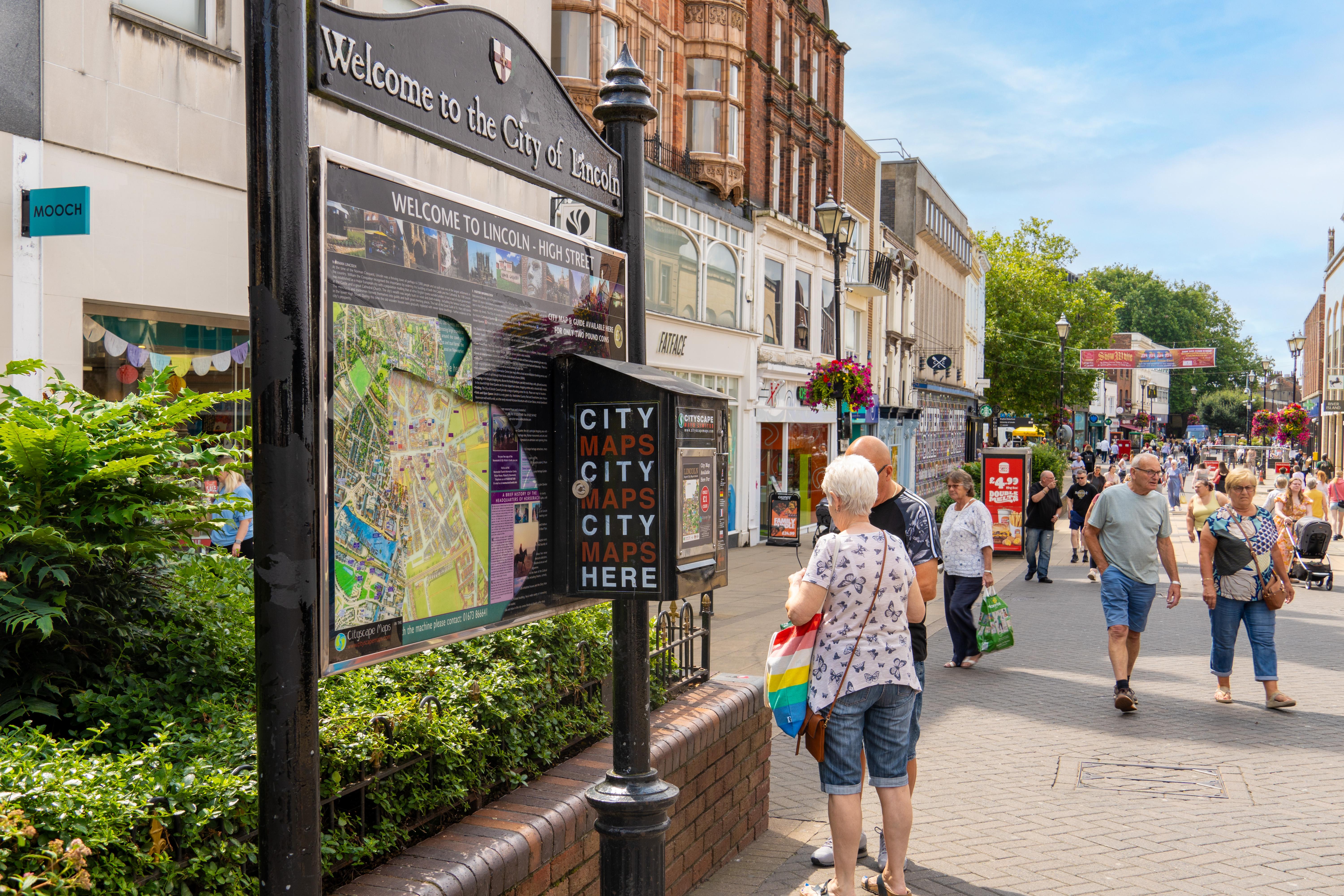 A City of Lincoln sign and map on a black stand in the centre of the High Street on a sunny day with people walking through town.