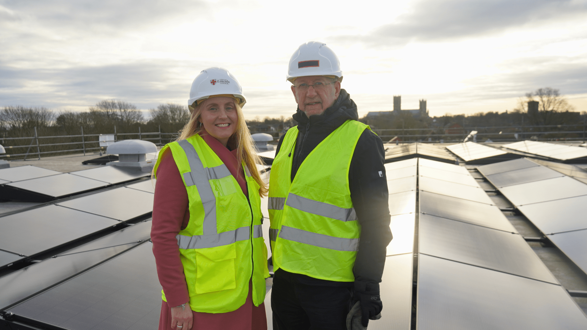 Two people standing on a roof wearing hard hats, with solar panels in the background
