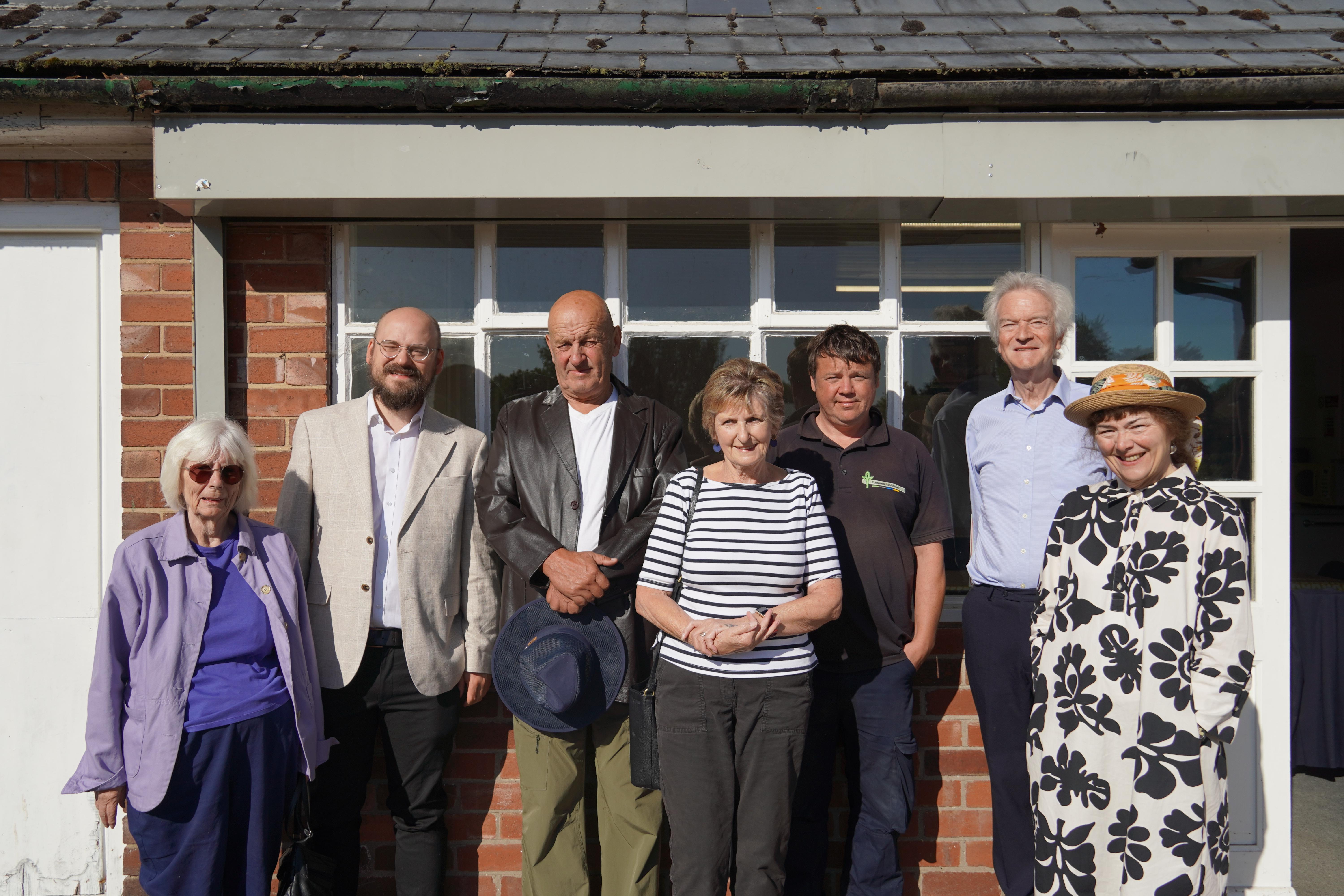 Three elderly women and four men of varying ages stood in a row in front of the window of a building.