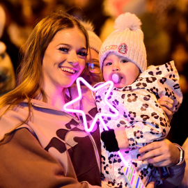 A woman holds a baby and a purple glowing star, the woman is smiling and they're both wrapped up warm at a Christmas Lights Switch On.
