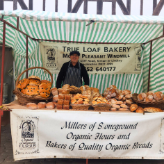 A smiling man in an apron stands behind his market stall on Castle Square, with lots of fresh, organic bread on display.