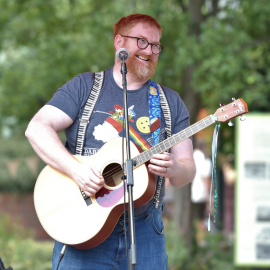 A man smile said song playing an acoustic guitar, with green trees in the background.