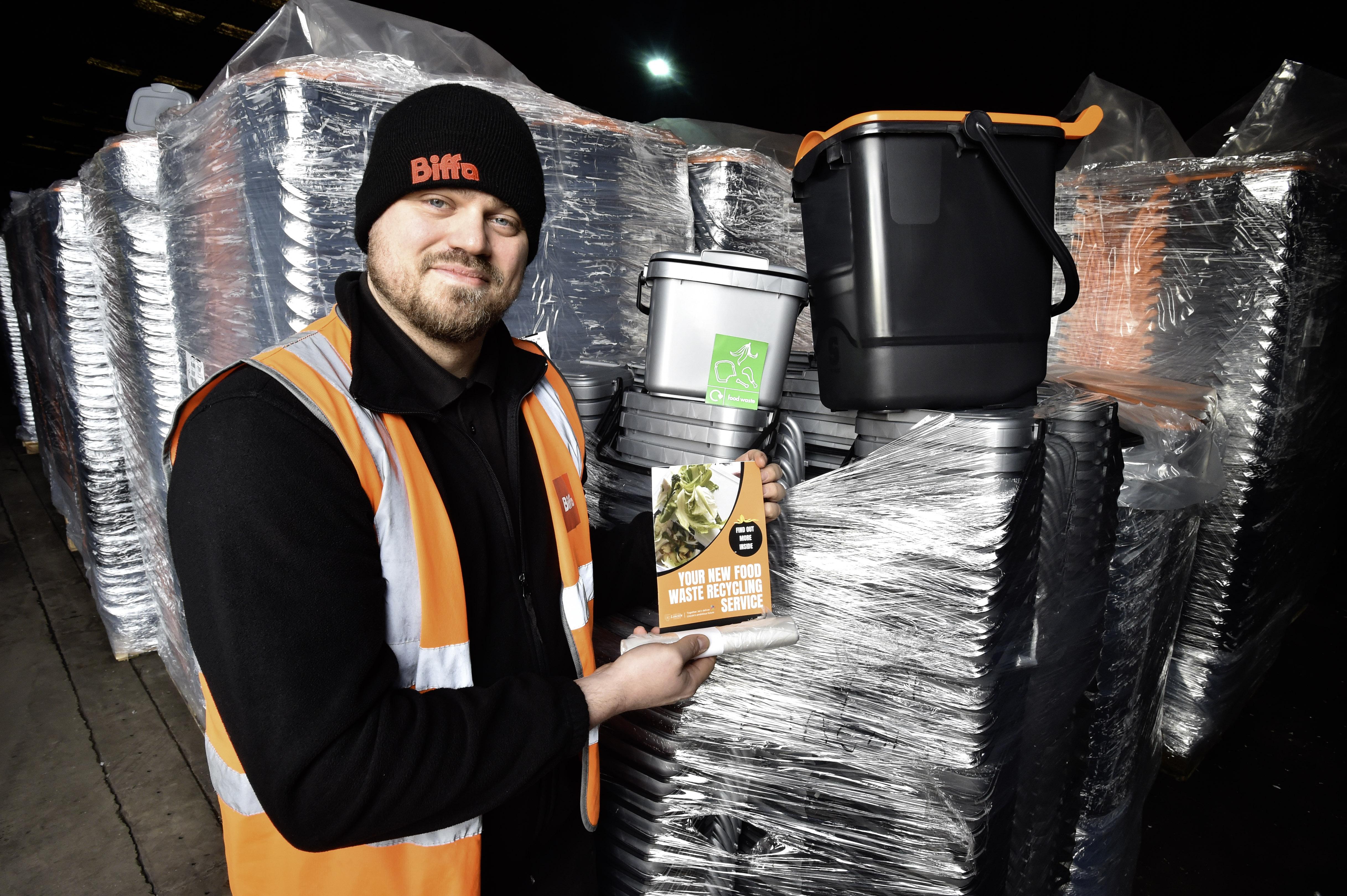 A white man in an orange hi-vis jacket wearing a black Biffa beanie holding a grey caddie, leaflet and roll of liners with the outdoor caddy on top of wrapped caddies behind him.