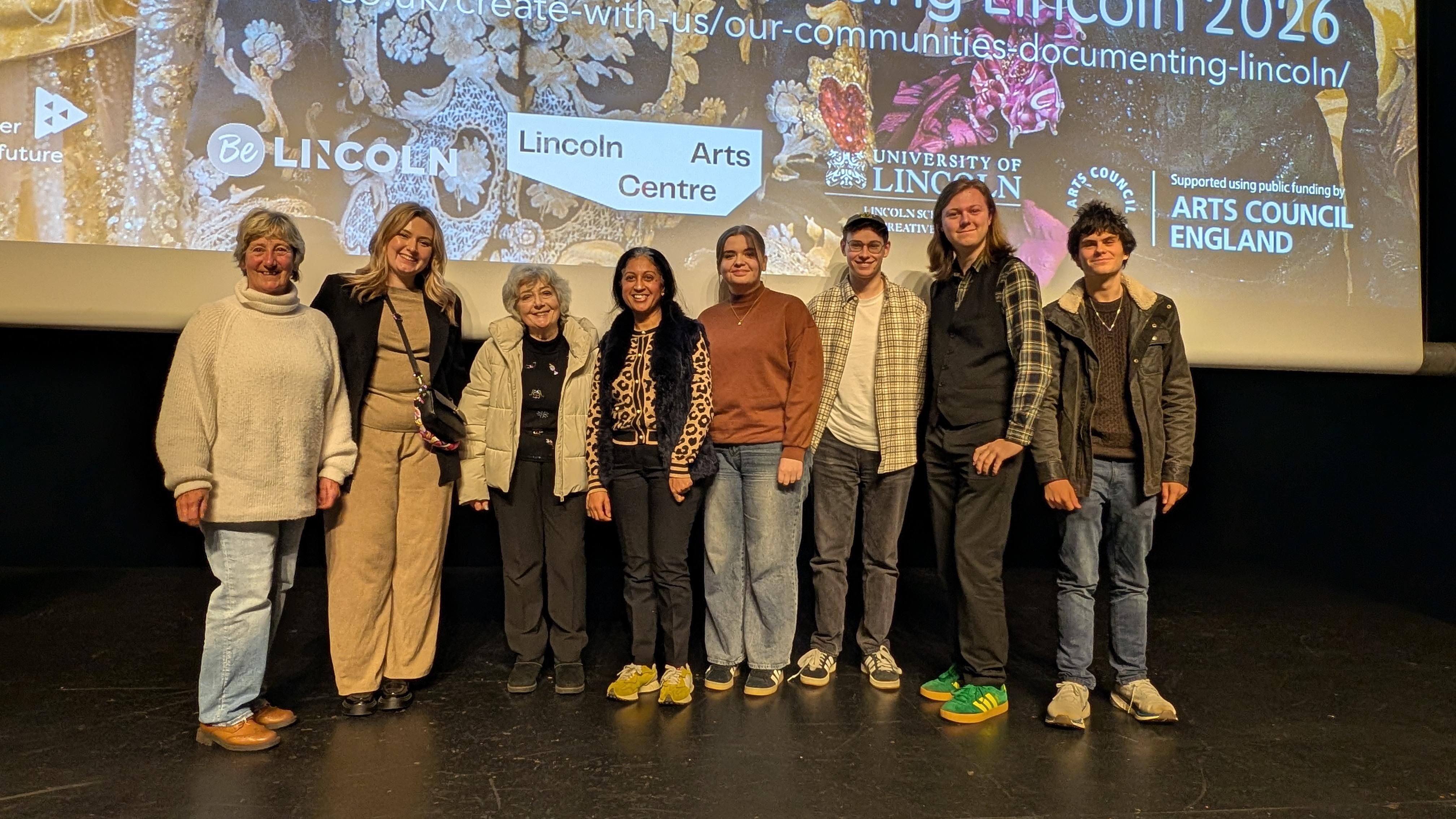 Eight people stood on a stage in a row in front of a large screen.
