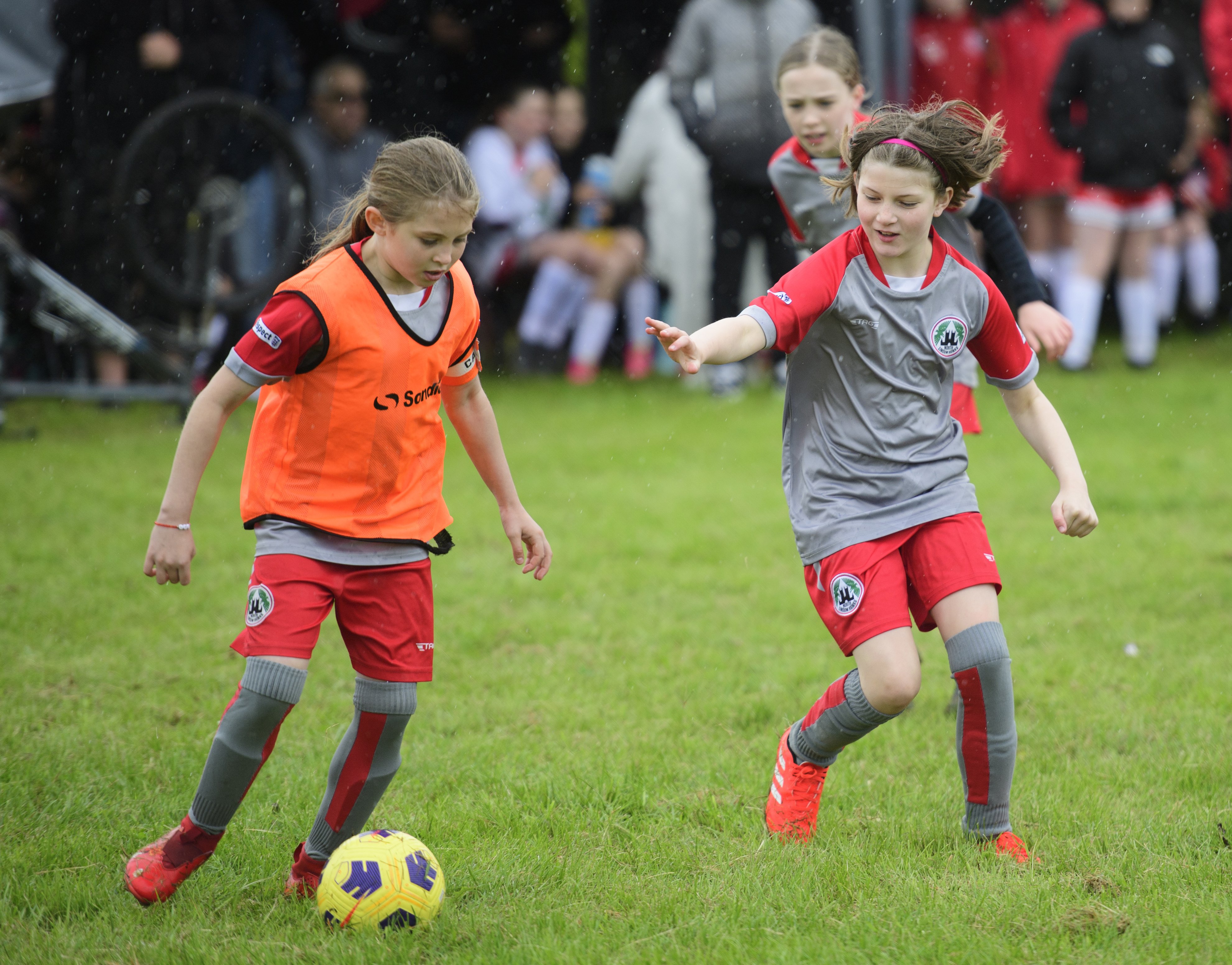 Two young children playing football on a field, one is wearing a bright orange bib.