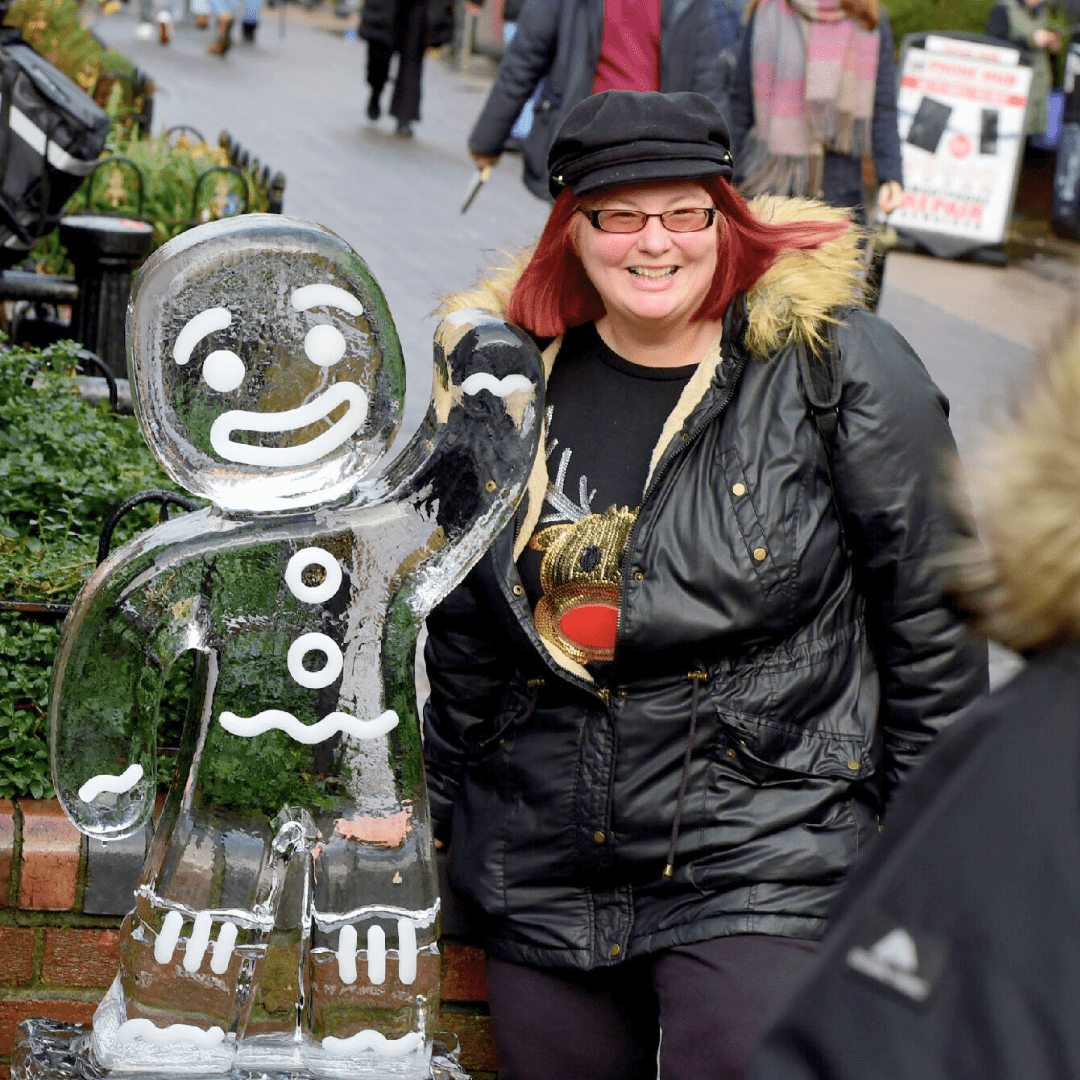 A woman in a black coat stands smiling next to an ice sculpture carved into the shape of a waving gingerbread man