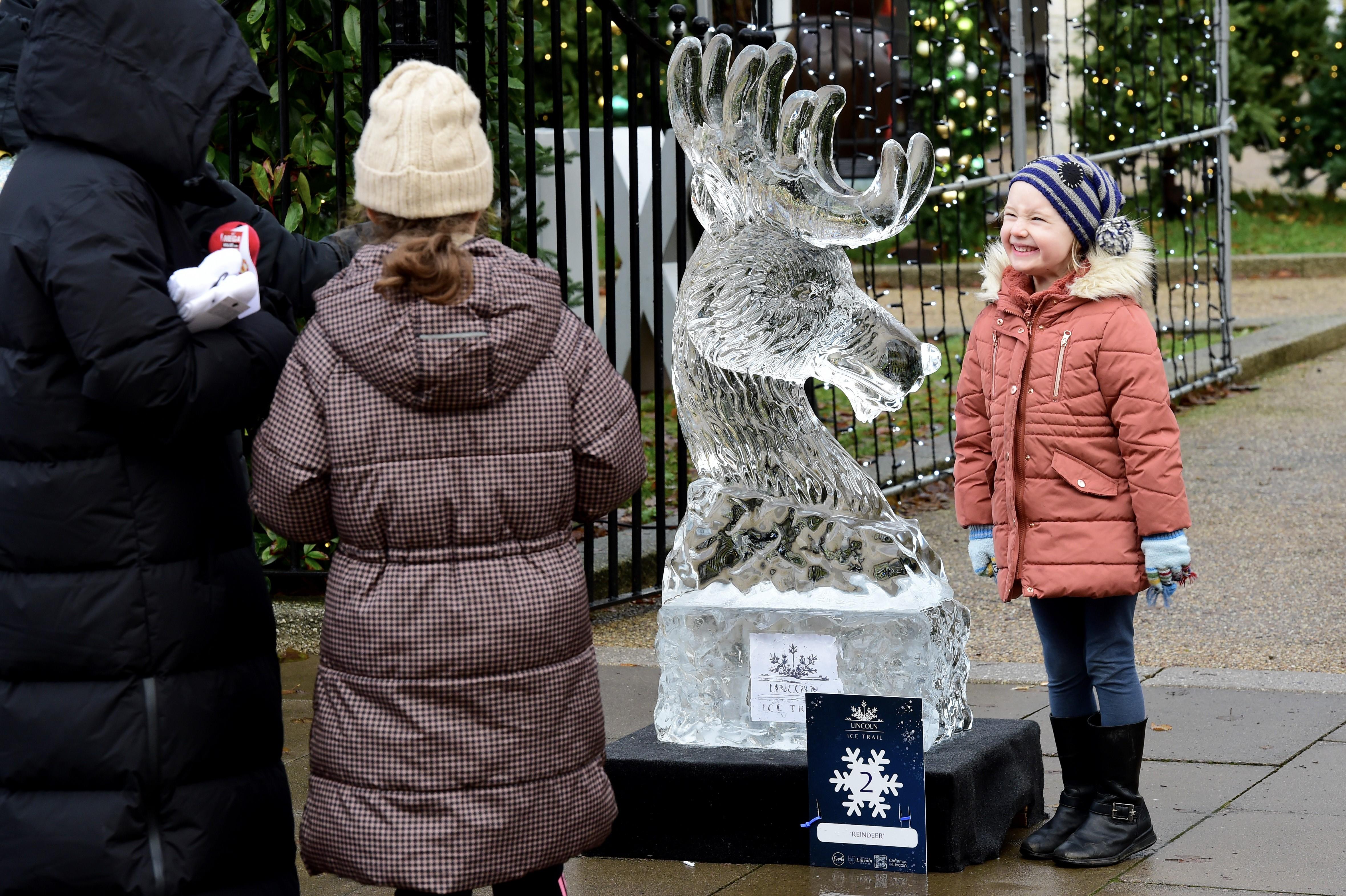 A little girl in coat, gloves and hat stands smiling for a photo next to an ice sculpture of a reindeer.