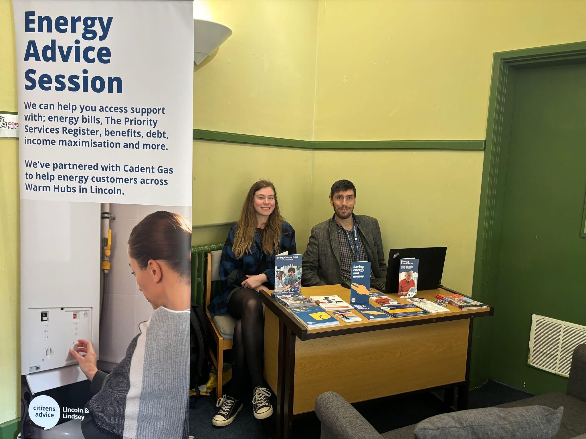 A young woman and man sat behind a desk smiling with an Energy Advice Session poster next to them.