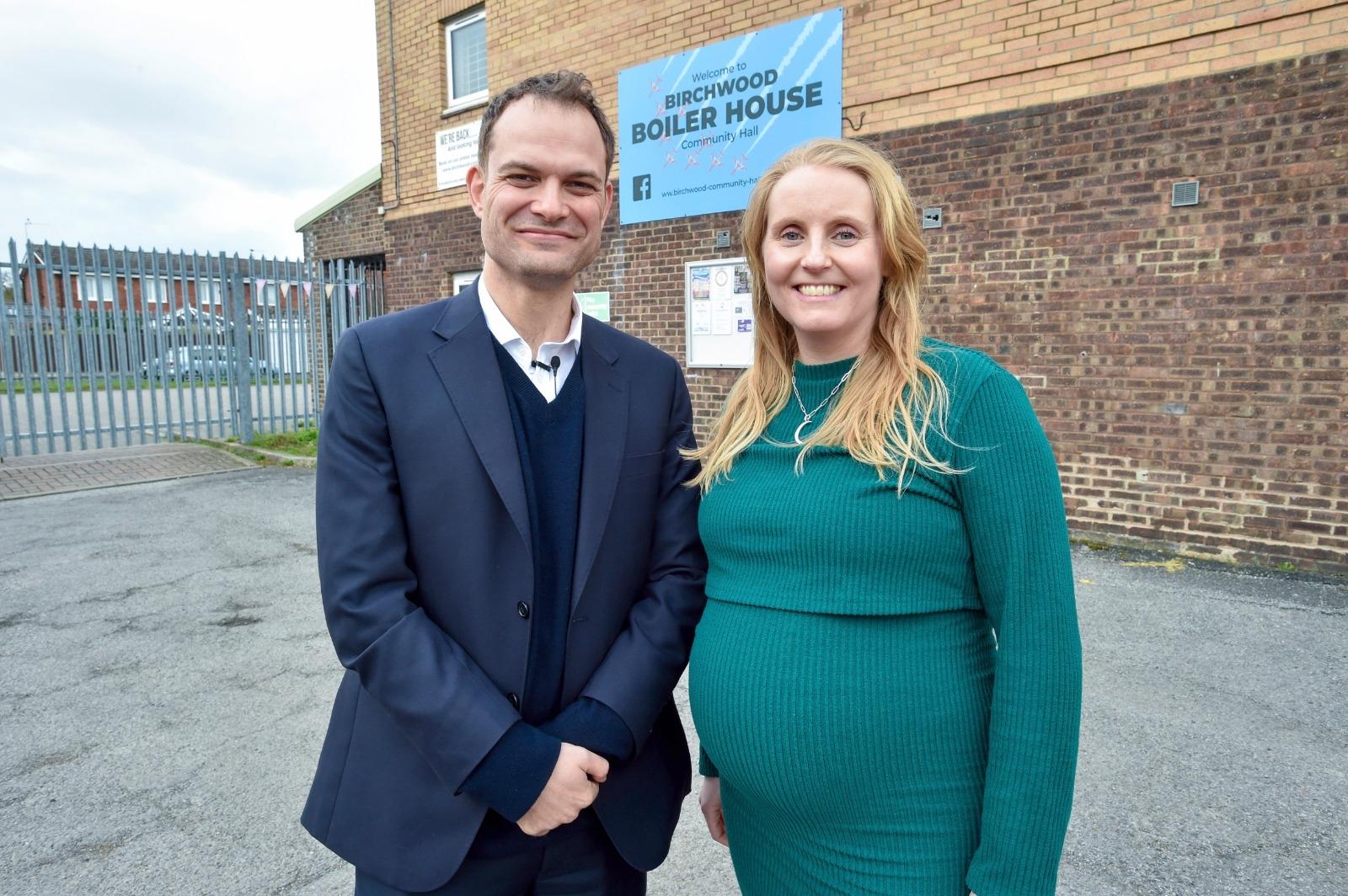A white man in a suit next to a pregnant white woman in a green dress outside a community centre.