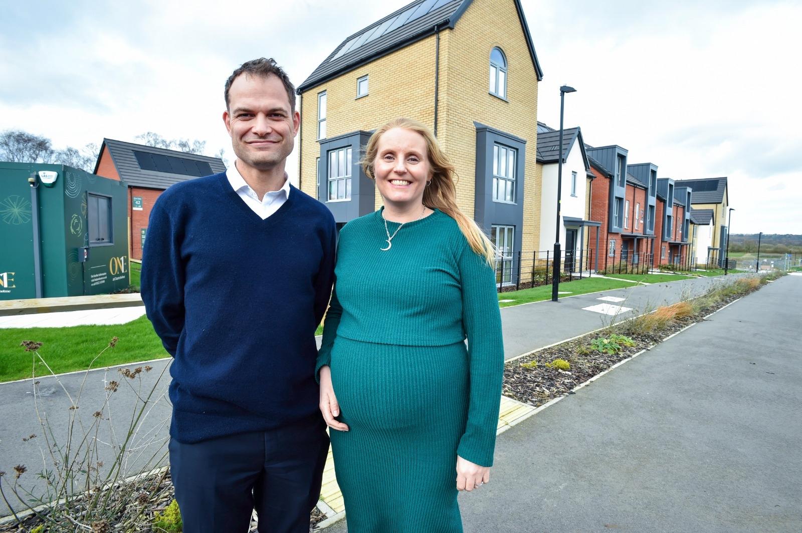 A white man and woman dressed smartly smiling for a photograph in front of new build houses.
