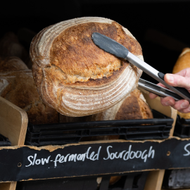 A pair of tongs pick up a fresh loaf of bread from a basket. The basket has a chalkboard sign on it saying 'slow fermented sourdough'.