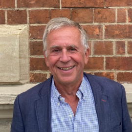 John stands against a brick wall and smiles at the camera in a blue shirt and blazer.