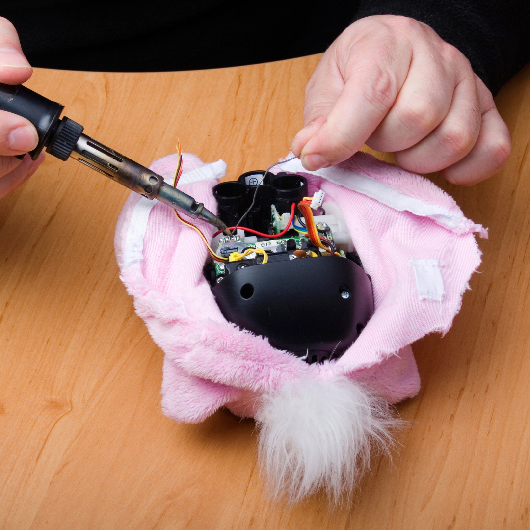 Pink fluffy toy unzipped to show battery pack, with somebody trying to repair the wires inside.