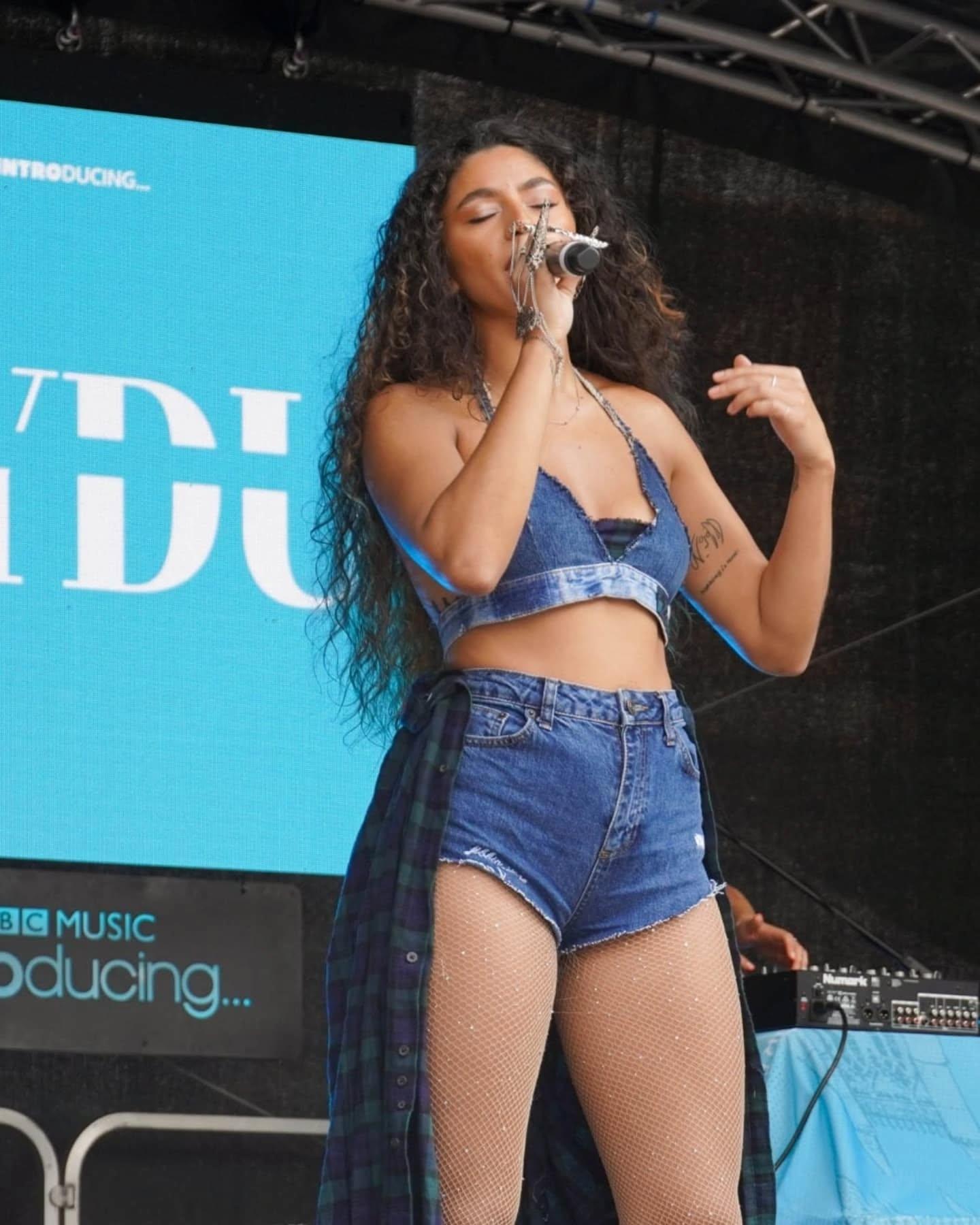 A young female on stage in a denim shorts and crop top co-ord singing into a microphone.