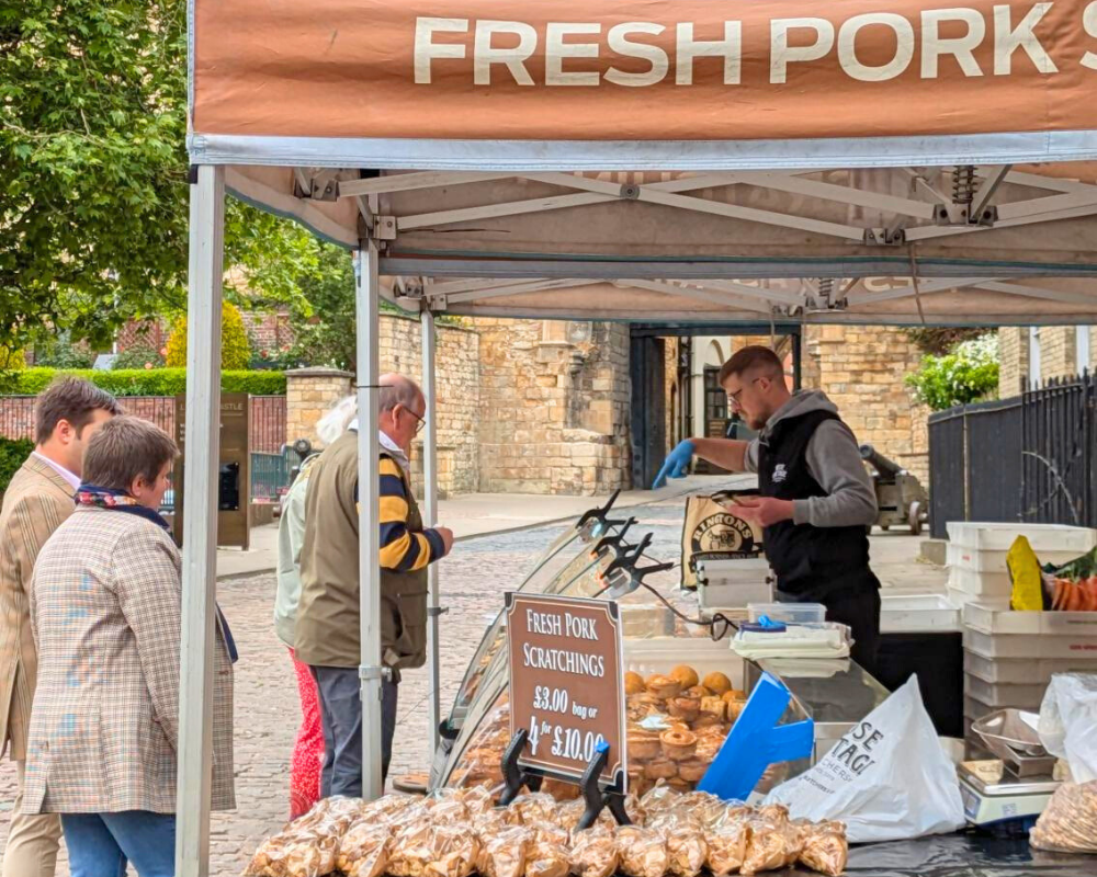 A few customers buying produce at the Fresh Pork Scratchings stall at the Lincoln Farmers' Market, with a green tree and Lincoln Castle in the background.