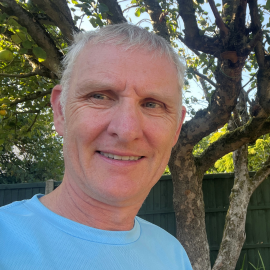 Nick wears a blue t-shirt standing in a garden in front of a tree in the sun, he smiles at the camera.