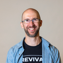 Pete wears a black t-shirt and blue shirt over, smiling at the camera.