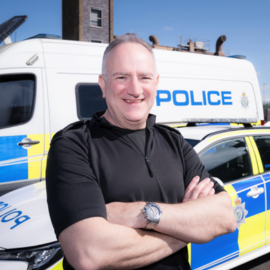 Phillip stands in front of a police van with his arms crossed, smiling at the camera.