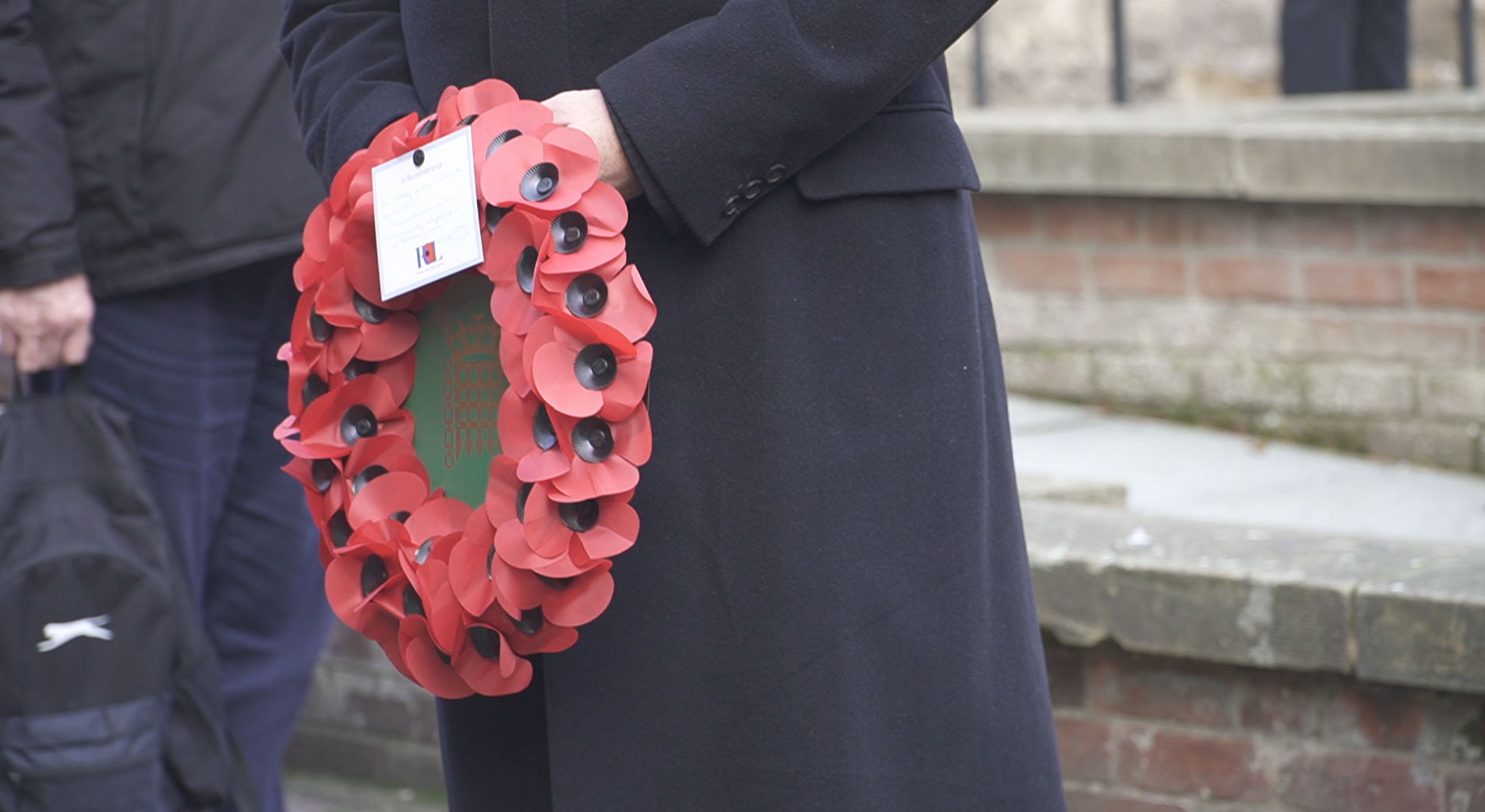 A person in a black coat that can only be seen from waist down holding a wreath of red poppy flowers.