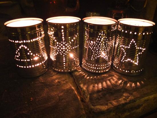 Four tin cans have been decorated with holes punched into them. They are in various star shaped patterns. Candles have been placed into each one and they are lighting up in the dark.