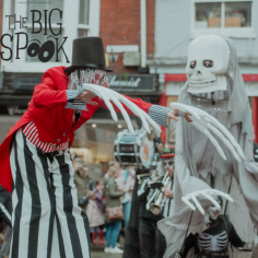 A spooky character wearing pinstripe trousers, a red jacket, and a top hat stands on stilts with huge skelton hands, next to a giant skeleton. A snapshot of the Strolling Bones parade in the Bailgate area, Lincoln.
