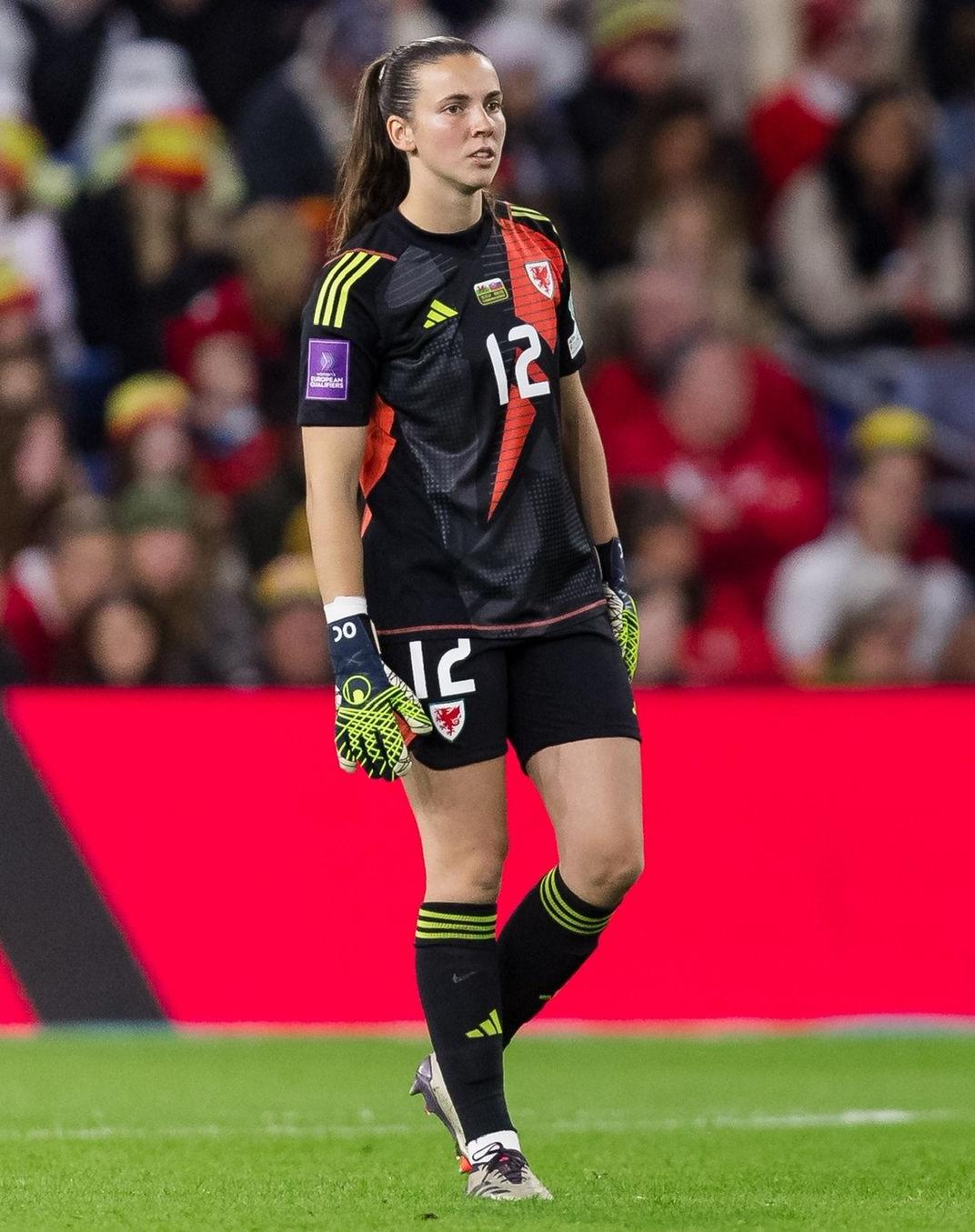 A female footballer in kit with her goalkeeper gloves standing on a pitch in a stadium.