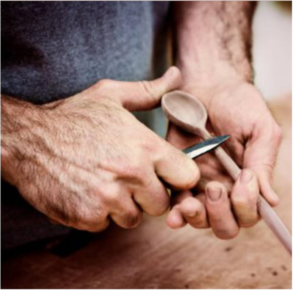 Zoomed in photo of a man holding a wooden spoon and whittling it with a knife.