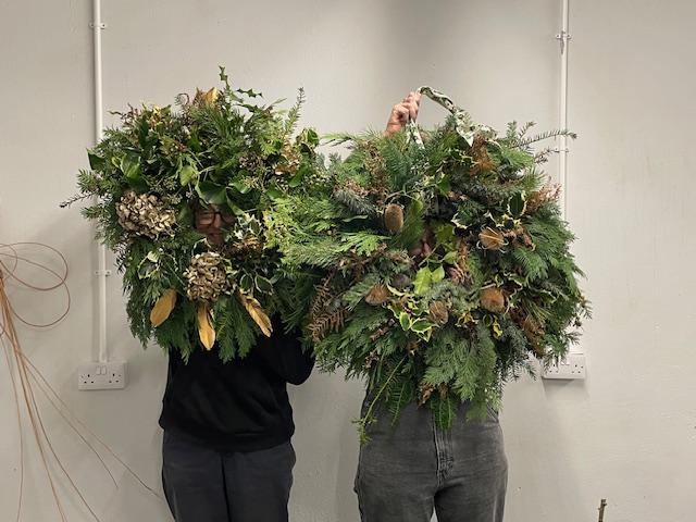 Two people holding up handcrafted wreaths in front of their faces.